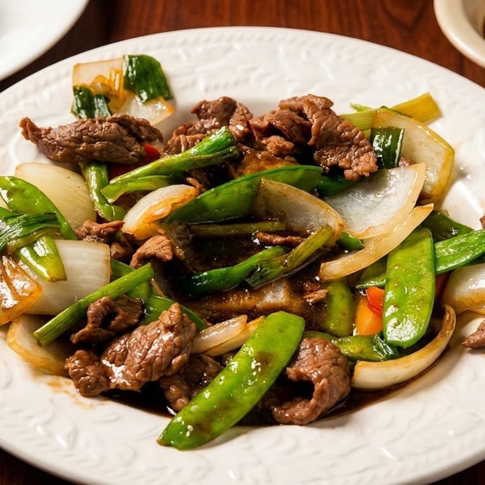 Beef stir-fry with snow peas, onions, scallions, and peppers served on a white plate with rice and dipping sauce.
