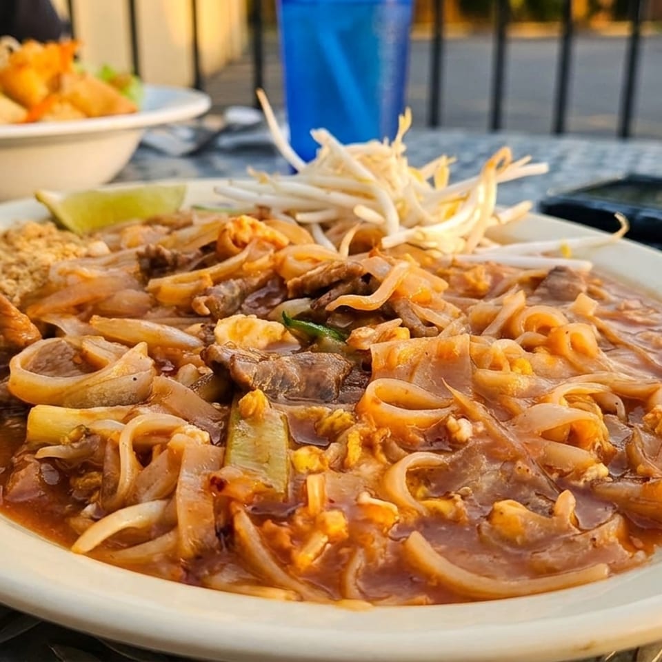 Stir-fried noodles with beef, egg, vegetables, bean sprouts, lime, and crushed peanuts served on an outdoor restaurant table.