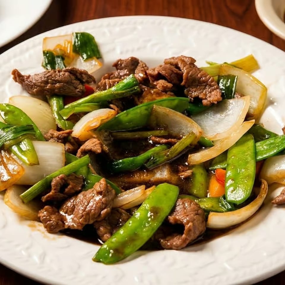 Stir-fried beef with snap peas, onions, carrots, and greens on a white plate, served with rice and dipping sauce.